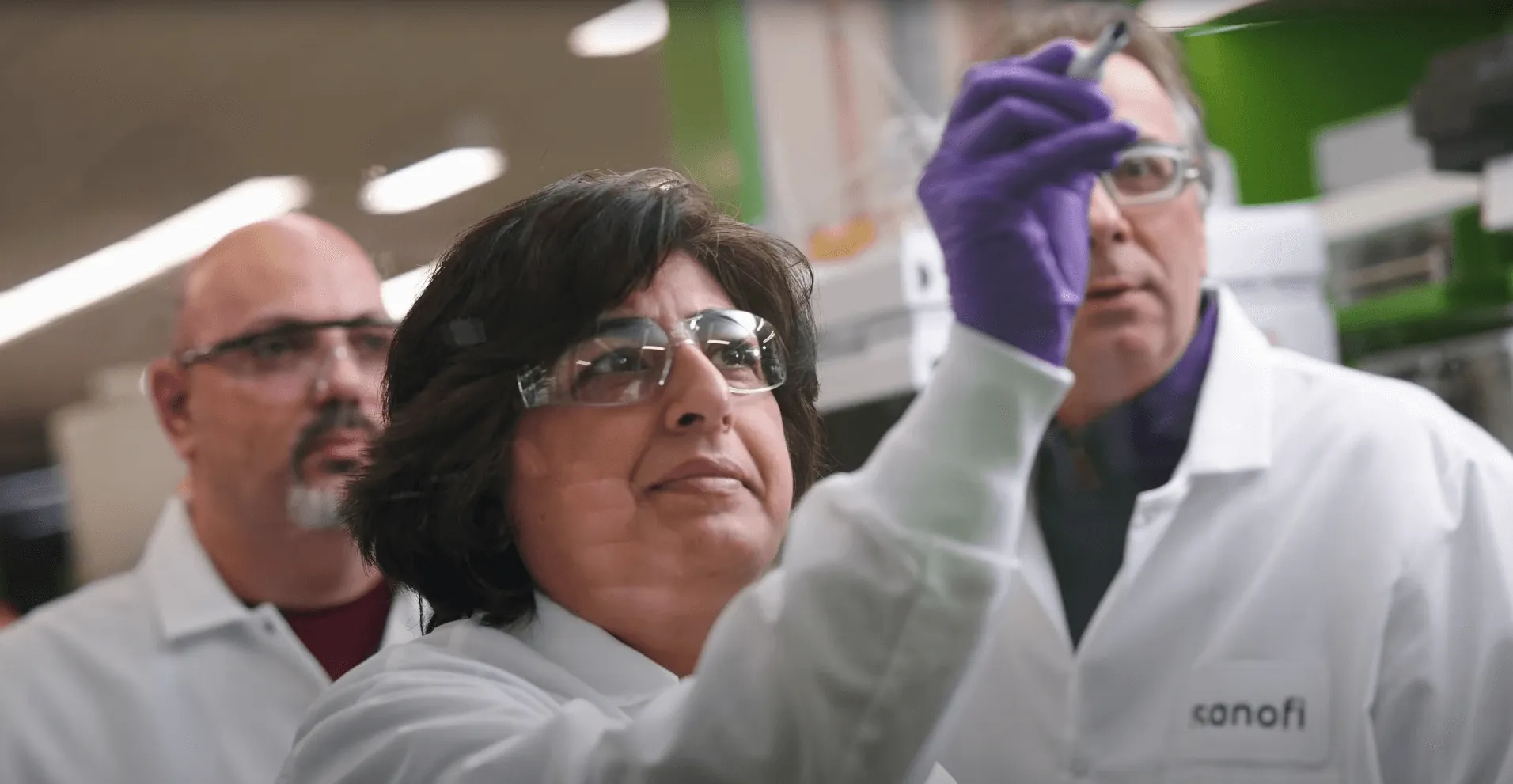 A scientist in a lab coat and safety glasses holds a sample up to the light, with two colleagues watching behind her.