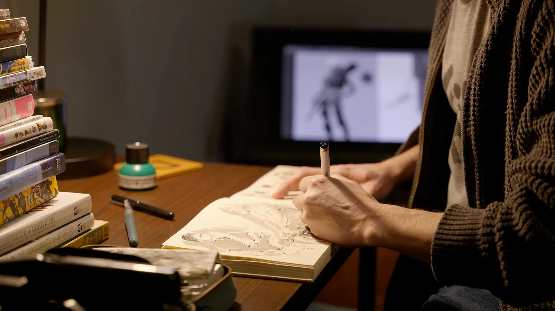 Hands sketching a character in a notebook at a lamplit desk, with concept art on the monitor behind.